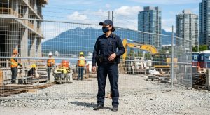 Professional security guard in uniform at Vancouver construction site, holding radio, vigilant amid fencing and equipment with city and mountain backdrop.